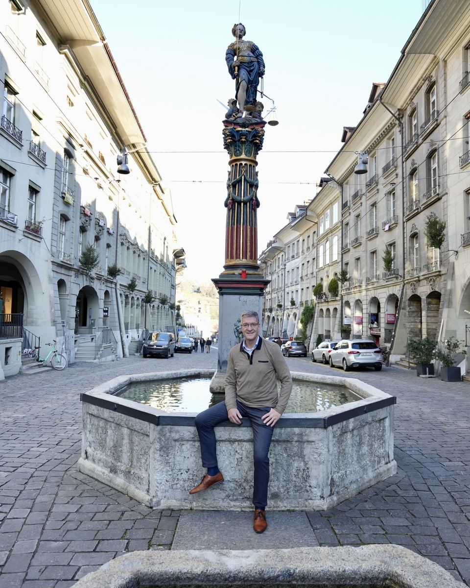 Arnold Frehner sitzt am Rand des Gerechtigkeitsbrunnens in der Berner Altstadt, darüber die Justitia-Figur.